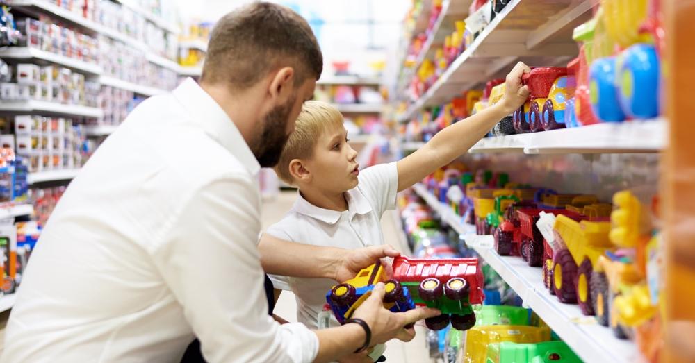A father and his son in the toy aisle of a store. The father is holding a plastic toy truck as his son looks on the shelf.