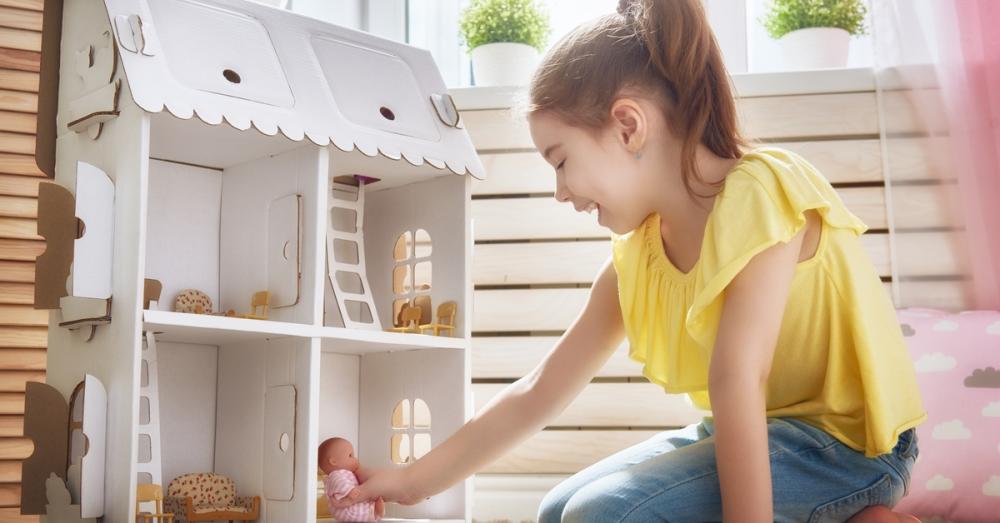 A little girl playing with a baby doll and a white cardboard doll house. She is sitting on a carpeted floor with a smile.