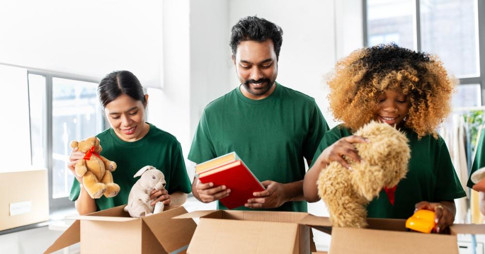 Three people sorting toys and books into cardboard boxes sitting on a table. They are wearing plain green t-shirts.