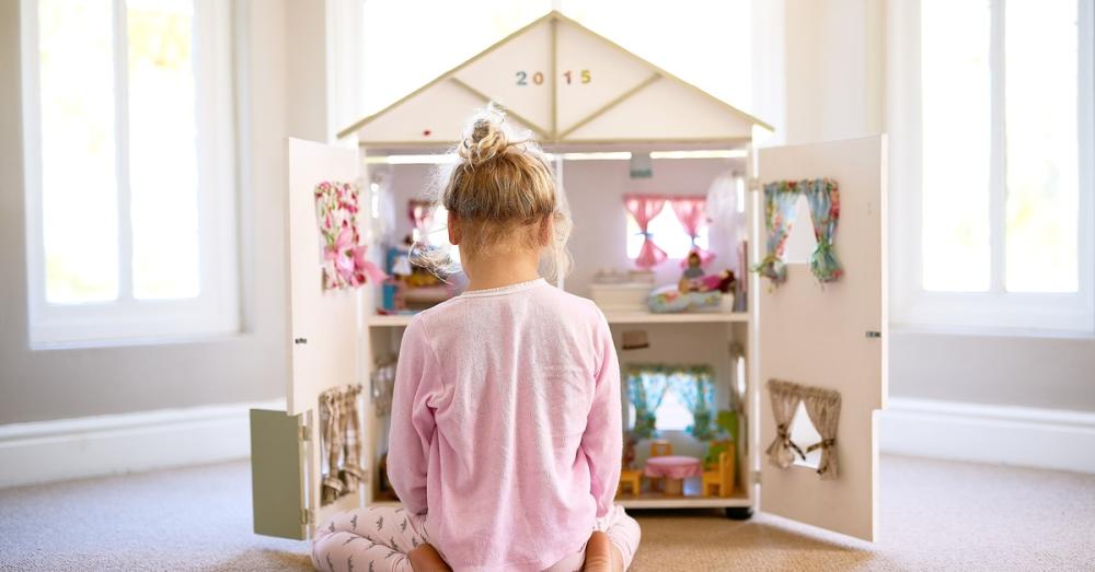 A little girl sitting in front of a doll house. The doll house has four sections within it with small figurines inside.