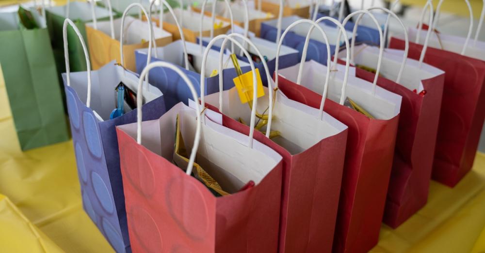 Green, yellow, blue, and red goodie bags lined up on the table with a yellow tablecloth. They are filled with items.