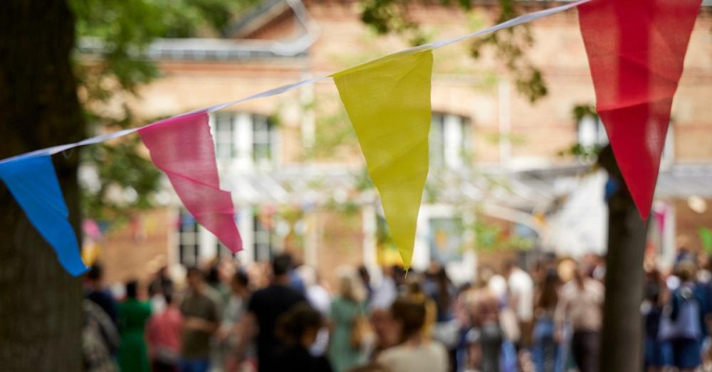 A decorative triangle banner hung up for an outdoor event. There is a bustling crowd of people in the background.