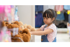 A little girl placing her hands on plushies inside of a retail store. She is wearing a pink shirt and overalls.