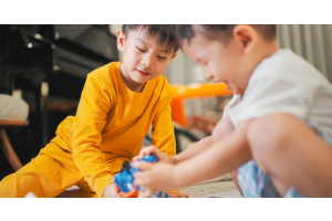Two young boys playing with action figures on the floor of their home. One of them holds a blue, bulky toy.