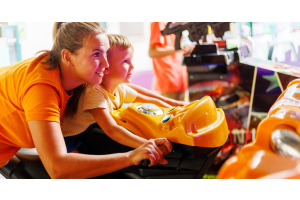 A mother and her son playing in an arcade. The son sits on a motorcycle machine while the mother controls the bike.