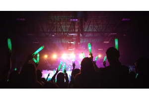 A large crowd of people at a dark indoor event. They are holding green light sticks in the air facing the main stage.