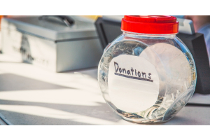 A container labeled “donations” filled with dollar bills and coins. It is sitting on a white table in front of metal boxes.