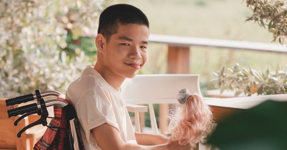 A boy sitting in a wheelchair on an outdoor patio. In his hands, he is holding a doll with bright pink hair.